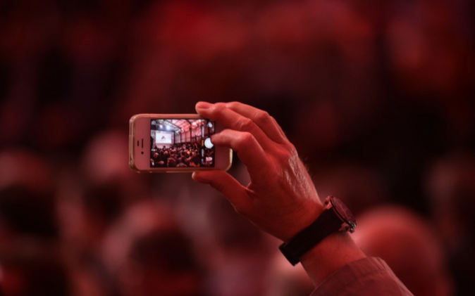 Eine Hand mit Handy, auf Bildschirm werden Feierlichkeiten aufgezeichnet.