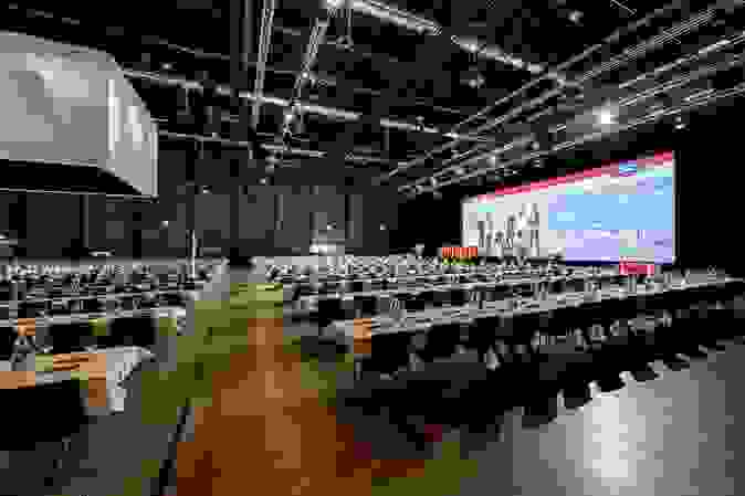 Leere Messehalle mit gedeckten Tischen und Stühlen. Im Hintergrund die Leinwand und das Podium