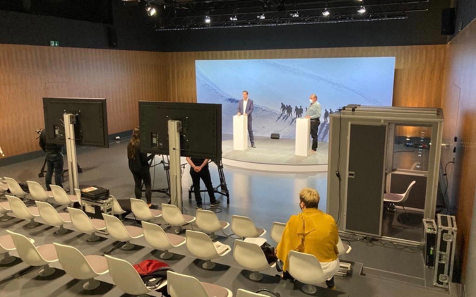 Zwei Personen auf Podium mit grosser Leinwand im Hintergrund. Vordergrund grosser Bildschirm von hinten. Aufzeichnung der digitalen Mitarbeiterinformation läuft. 