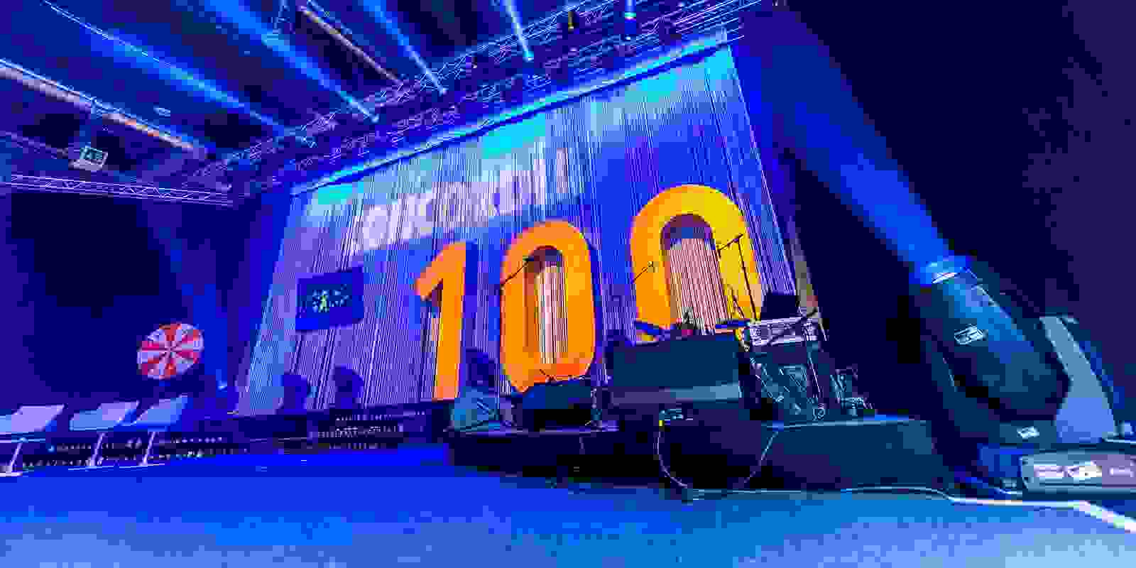 Bühne mit Logo-Projektion und Nummer 100. Im Vordergrund die Instrumente der Band. 