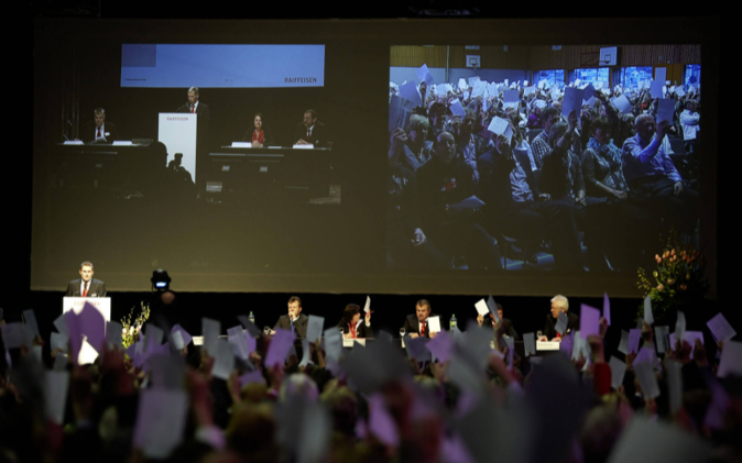 Leinwand mit Podium im Hintergrund, vorne Aktionäre bei einer Abstimmung. 