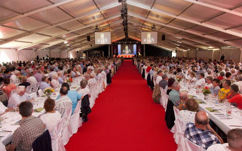 Zelt mit Gästen an den festlich geschmückten Tischen und Stühlen, Leinwand mit rotem Teppich. 