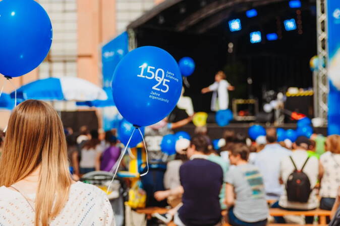ewl Jubiläum Im Vordergrund Person mit Ballon mit Aufschrift 150 Jahre Erfahrung, 25 Jahre Eigenständigkeit im Rahmen der Jubiläumsfeierlichkeiten von ewl. Im Hintergrund Tische und Gäste und eine Bühne.