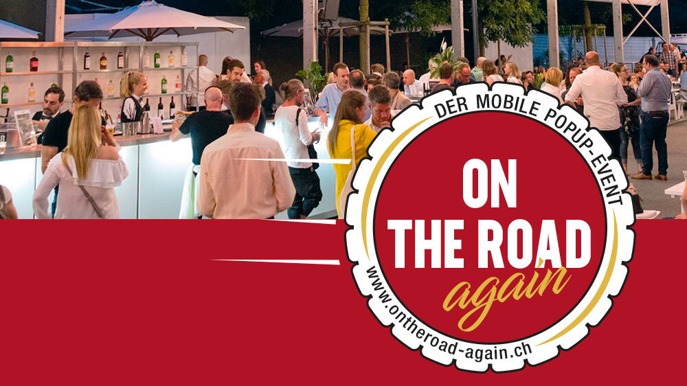Bar mit Gästen an einem Anlass, vorne roter Balken und Logo "on the Road again". 