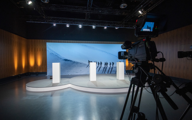 Digitale Mitarbeiterinformation Podium mit grosser Leinwand im Hintergrund. Vordergrund steht eine Kamera, die die digitale Mitarbeiterinformation aufzeichnet.