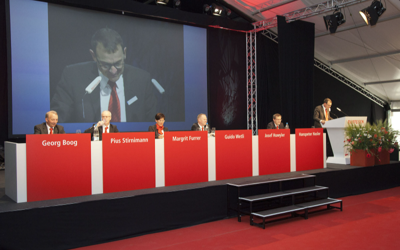 GVs und Delegiertenversammlungen der Raiffeisen schweizweit Das Podium mit den Redner und die Leinwand.