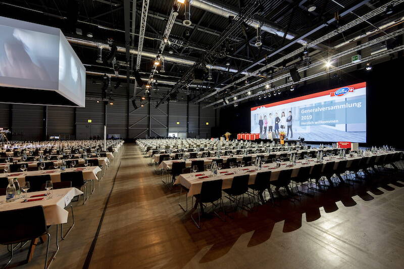 GV Emmi AG Leere Messehalle mit gedeckten Tischen und StĂŒhlen. Im Hintergrund die Leinwand und das Podium