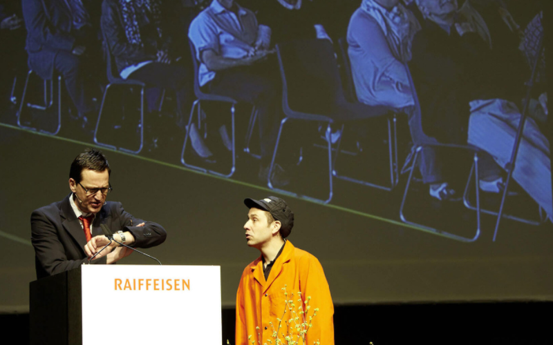 GVs und Delegiertenversammlungen der Raiffeisen schweizweit Komiker auf dem Podium während einer Generalversammlung.