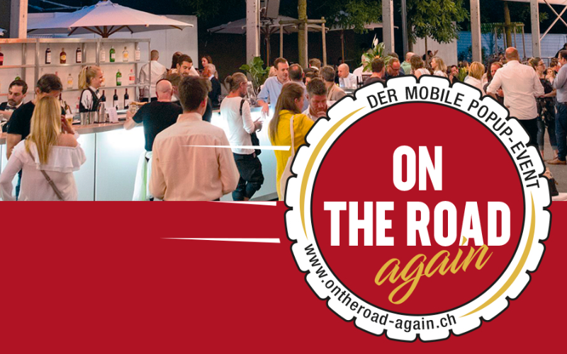 Bar mit Gästen an einem Anlass, vorne roter Balken und Logo "on the Road again". 