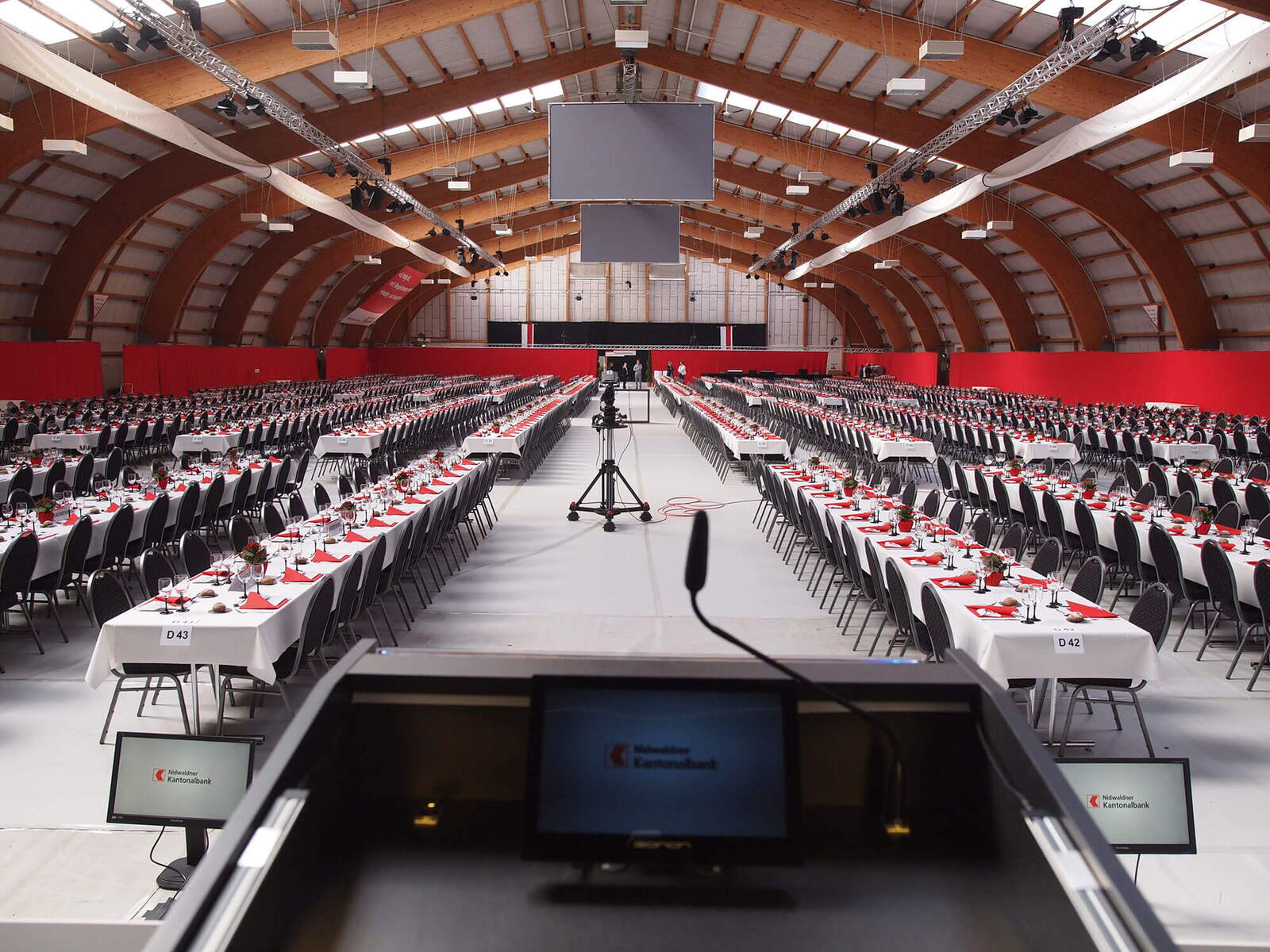 Partizipations-Versammlung der Nidwaldner Kantonalbank Eingerichtete Tennishalle mit gedeckten Tischen. Blick vom Rednerpult aus zu den Gästen. Kleiner Bildschim auf Rednerpult im Vordergrund.