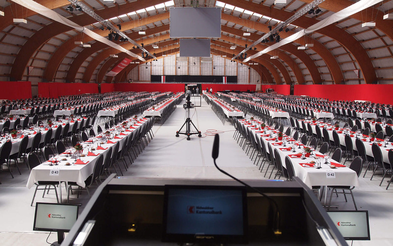 Partizipations-Versammlung der Nidwaldner Kantonalbank Eingerichtete Tennishalle mit gedeckten Tischen. Blick vom Rednerpult aus zu den Gästen. Kleiner Bildschim auf Rednerpult im Vordergrund.